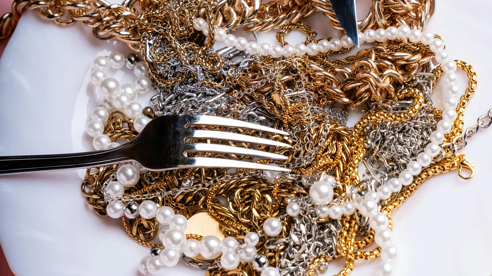 Tangled gold and silver chains and pearl necklaces piled on a white plate with a fork and knife on pink fabric
