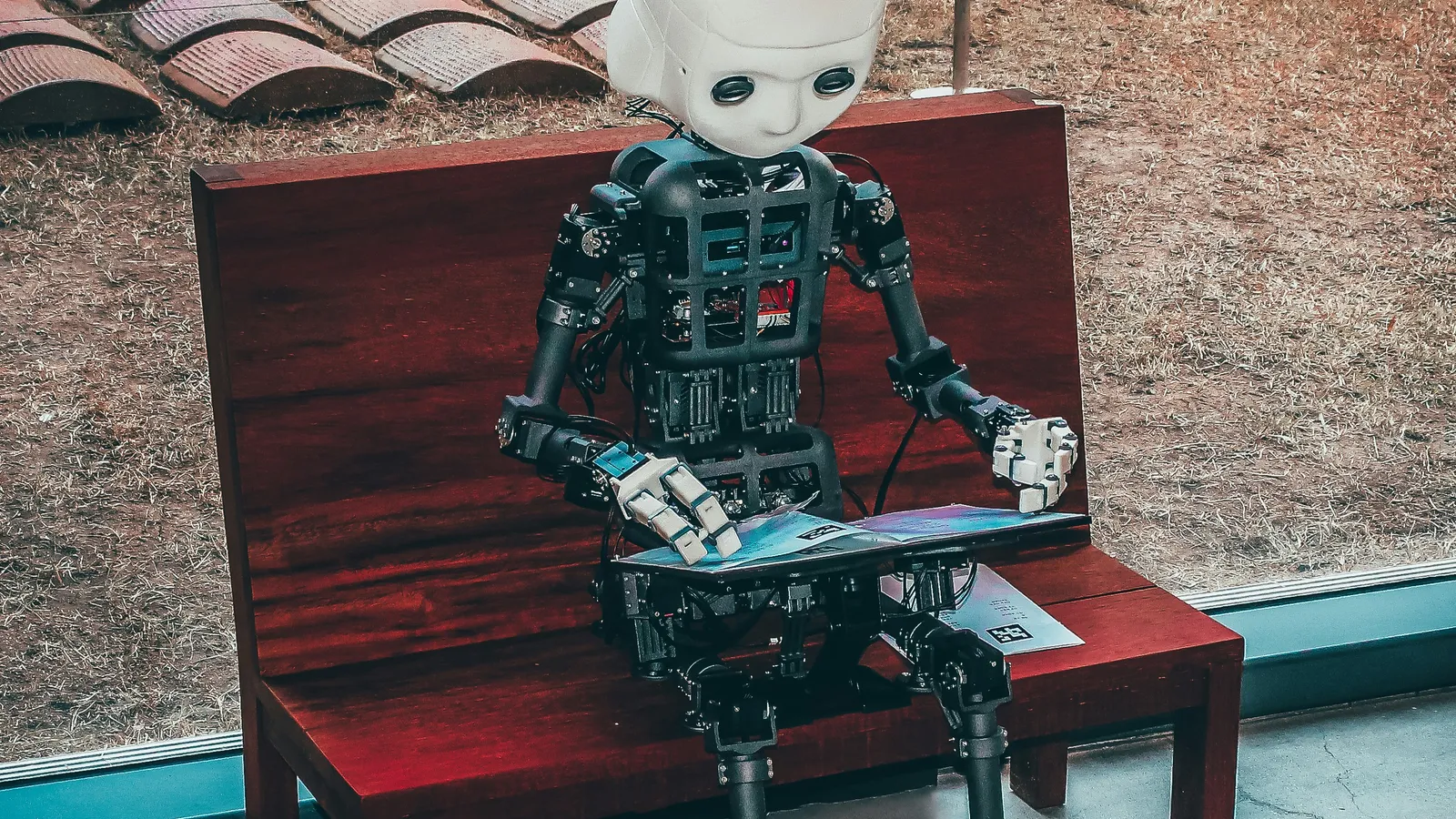 Small humanoid robot with a white head sits on a red wooden bench, reading a tablet beside a large window