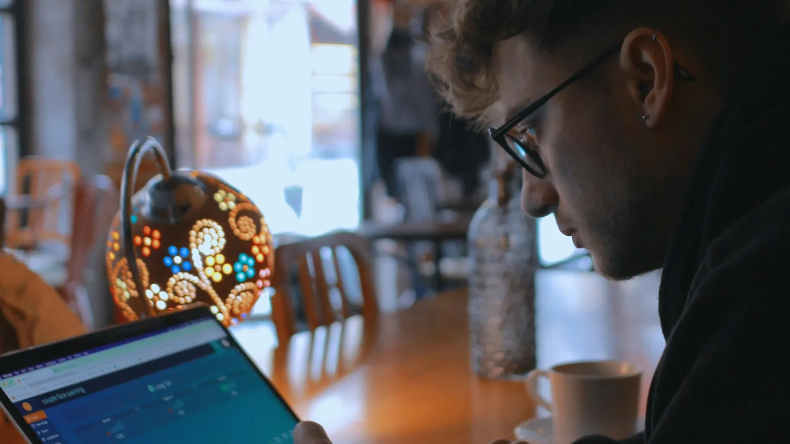 Man in glasses and a dark hoodie works on a laptop showing a teal analytics dashboard at a wooden cafe table