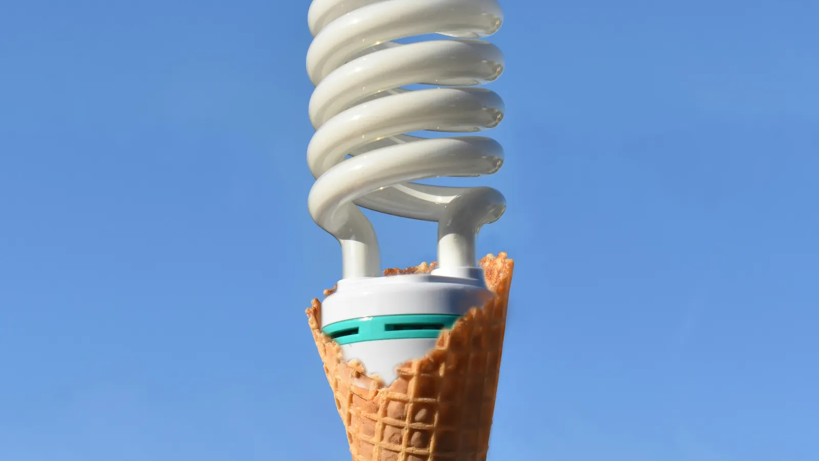 Hand holding a waffle ice cream cone topped with a spiral compact fluorescent light bulb against a clear blue sky