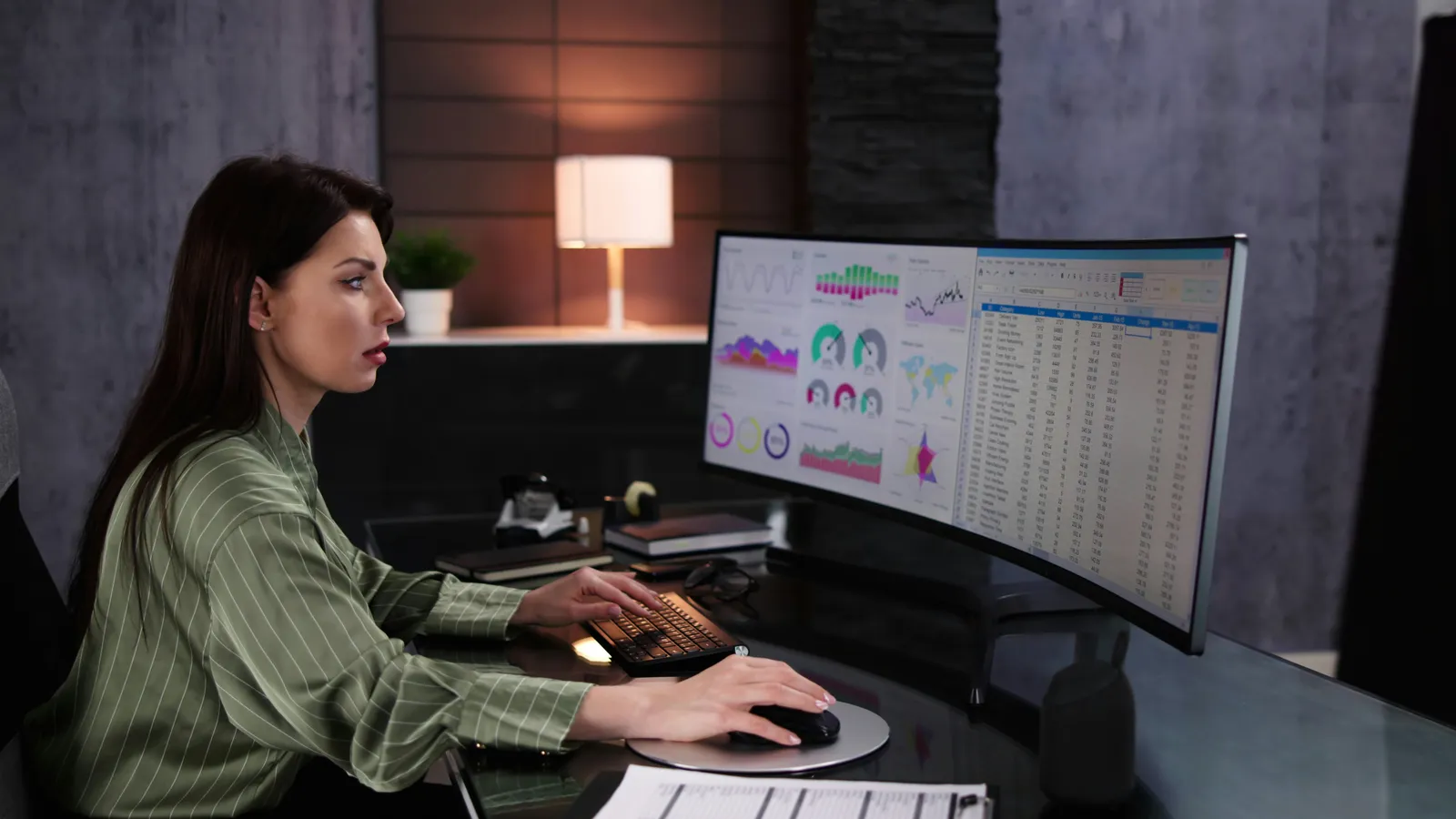 Woman in a green striped blouse works at a desk, studying analytics dashboards and a spreadsheet on a curved ultrawide monitor