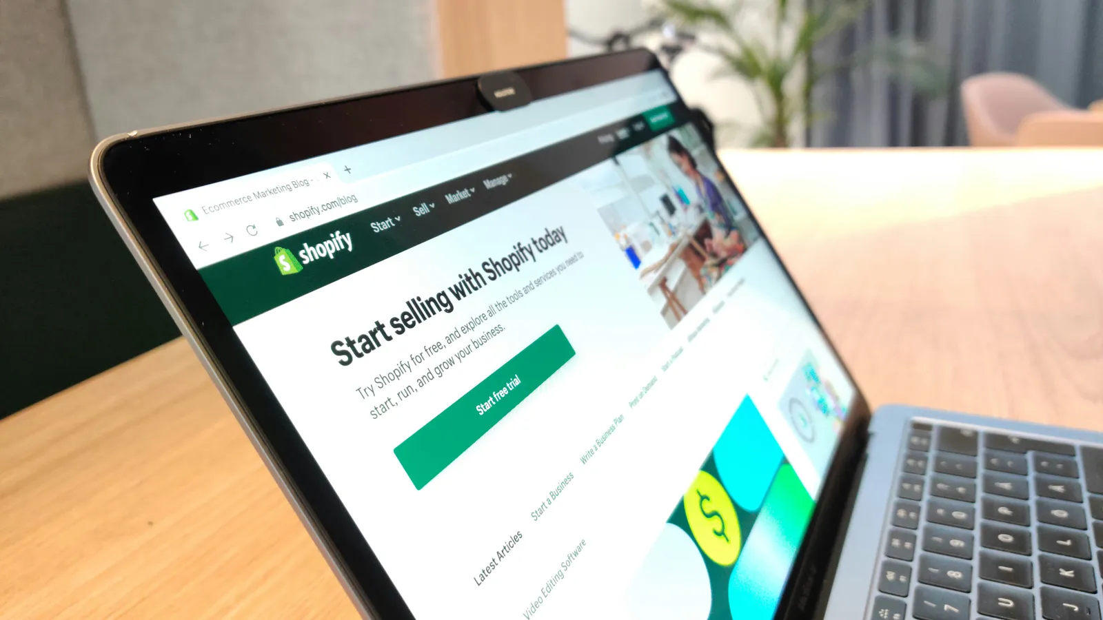 Laptop on a wooden desk showing the Shopify homepage with a Start free trial button, in a bright modern office