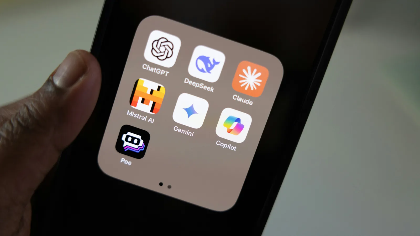Hand holding a smartphone showing an AI folder with ChatGPT, DeepSeek, Claude, Mistral AI, Gemini, Copilot and Poe app icons