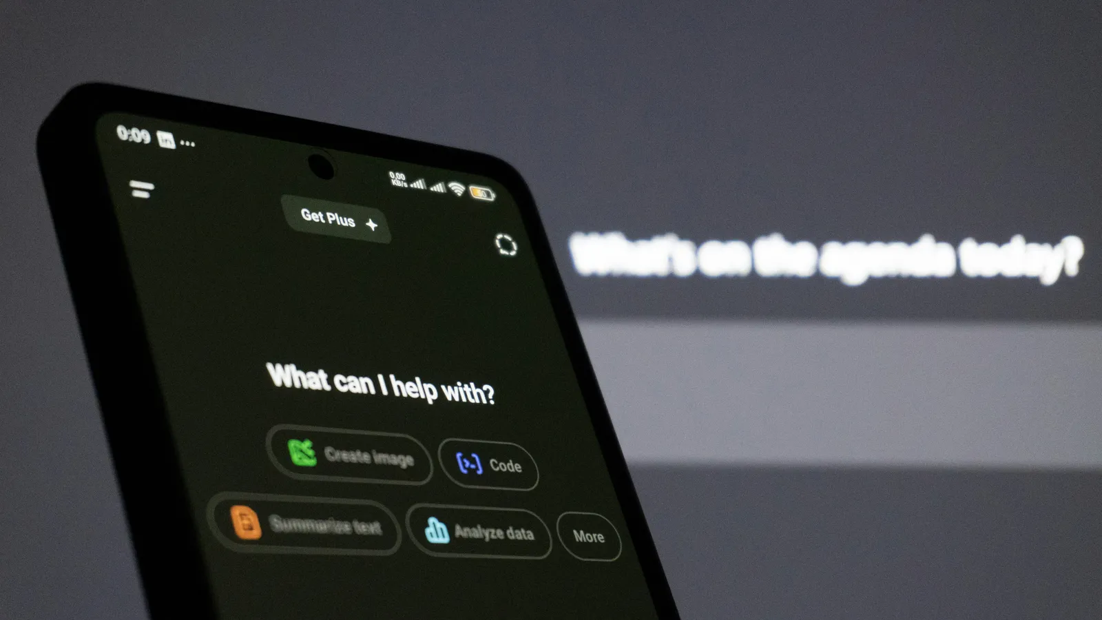 Smartphone showing the ChatGPT app with prompt suggestions, in front of a blurred screen reading What's on the agenda today?